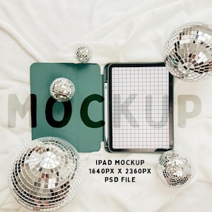 May include: A green iPad case with a white iPad inside. The iPad is displaying a grid pattern. The iPad is surrounded by silver disco balls on a white fabric background. The text "IPAD MOCKUP 1640PX X 2360PX PSD FILE" is below the iPad.