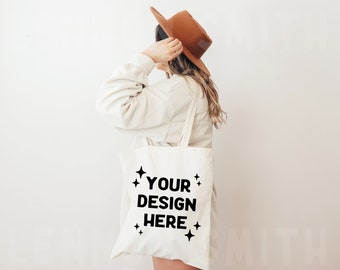 Puede incluir: Una bolsa de tela blanca con texto negro que dice "YOUR DESIGN HERE" y pequeñas estrellas alrededor del texto. La bolsa está siendo sostenida por una persona que lleva un suéter blanco y un sombrero marrón.