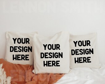 May include: Three white pillow covers with black text that reads "Your Design Here" on a white bed with a brown blanket.