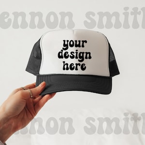 May include: A black and white trucker hat with the text "your design here" in a bold, black font. The hat has a white front panel, a black brim, and a black mesh back. The hat is held by a hand.
