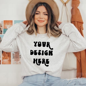 Può includere: Una felpa grigia a maniche lunghe e girocollo. La felpa ha la scritta "YOUR DESIGN HERE" stampata sul davanti in un font nero retrò.