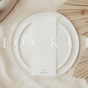 May include: A white rectangular card, labeled "4 x 9" and "PSD+JPG", rests on a white plate, with another plate beneath. The word "MOCKUP" is displayed in large, transparent letters. The background features a beige fabric and dried palm leaves.