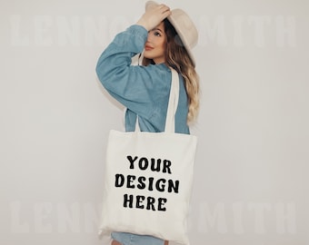 May include: A white tote bag with black text that says "YOUR DESIGN HERE". A person is holding the bag and wearing a blue denim jacket and a beige hat.