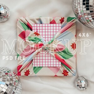 Op de afbeelding: Een kleurrijk cadeaupapier van stof met een witte kaart met een roze rasterpatroon. Het cadeaupapier is vastgebonden met een knoop en de kaart is gelabeld met "4x6". Het cadeaupapier is omringd door zilveren discoballen.