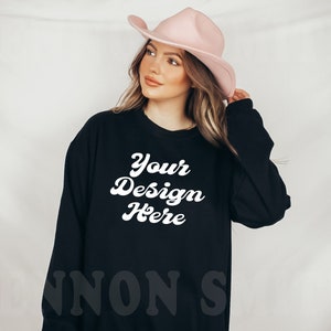 Puede incluir: Sudadera negra de manga larga con el texto "Your Design Here" en blanco en una fuente retro. La sudadera la lleva puesta una persona que lleva un sombrero vaquero rosa.
