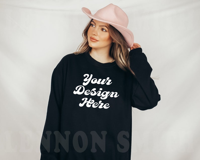 Puede incluir: Sudadera negra de manga larga con el texto "Your Design Here" en blanco en una fuente retro. La sudadera la lleva puesta una persona que lleva un sombrero vaquero rosa.