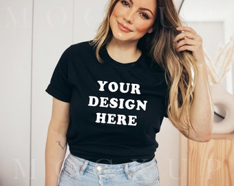May include: A woman wearing a black t-shirt with the text "YOUR DESIGN HERE" in white. She is smiling and has long blonde hair. She is wearing blue jeans.