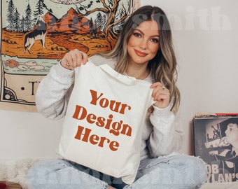 May include: A white tote bag with the text "Your Design Here" in orange letters. The bag is being held by a person wearing a grey sweater and blue jeans.