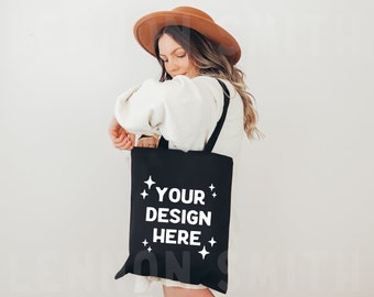 May include: A black tote bag with white text that says "YOUR DESIGN HERE" and stars. The bag is being held by a person wearing a white sweater and a brown hat.