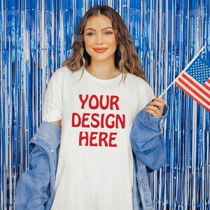 Puede incluir: Una camiseta blanca con el texto "YOUR DESIGN HERE" en letras rojas. La camiseta la lleva puesta una persona delante de un fondo de cortina de flecos azul y blanco. La persona sostiene una peque&ntilde;a bandera estadounidense.