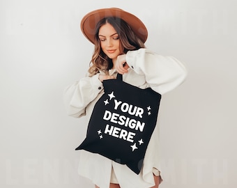 May include: A woman wearing a brown hat and a white shirt is holding a black tote bag with the text "YOUR DESIGN HERE" in white.