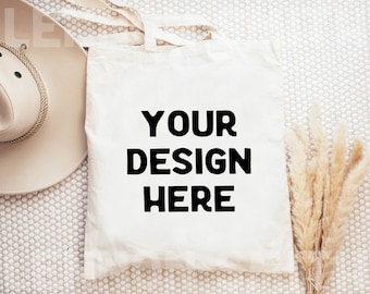 Puede incluir: Bolsa de tela blanca de lona con texto negro que dice "YOUR DESIGN HERE". La bolsa está tendida sobre una superficie blanca texturizada con un sombrero beige y hierba de la pampa seca.