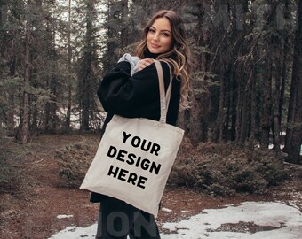 May include: A woman wearing a black jumper is holding a white tote bag with the text "YOUR DESIGN HERE" printed on it. She is standing in a snowy woodland.
