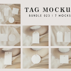 Puede incluir: Un conjunto de siete etiquetas de regalo blancas en blanco con cuerda atada. Las etiquetas están dispuestas sobre un fondo de tela beige con una superficie de madera debajo. El texto "TAG MOCKUP BUNDLE 023 | 7 MOCKS" se muestra en la parte superior de la imagen.