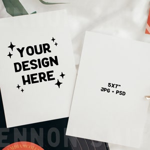 Op de afbeelding: Een witte kaart met de tekst "YOUR DESIGN HERE" in zwarte letters en kleine sterren rond de tekst. De kaart ligt op een zwarte vinylplaat met het woord "LENNON" zichtbaar. Een tweede witte kaart bevindt zich rechts van de eerste kaart met de tekst "5X7" JPG + PSD in zwarte letters.