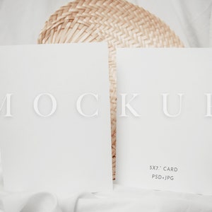 Op de afbeelding: Twee witte 12,7 x 17,8 cm kaart mockups met het woord "MOCKUP" in een licht lettertype. De kaarten liggen op een witte stoffen ondergrond met een geweven waaier op de achtergrond. De tekst "5x7" CARD PSD+JPG" staat op de rechterkaart.