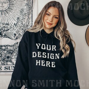 Pu&ograve; includere: Una felpa con cappuccio nera con il testo "YOUR DESIGN HERE" stampato in bianco sul davanti. La modella indossa la felpa e ha i capelli lunghi e biondi.