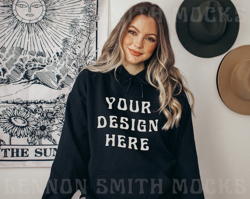 Pu&ograve; includere: Una felpa con cappuccio nera con il testo "YOUR DESIGN HERE" stampato in bianco sul davanti. La modella indossa la felpa e ha i capelli lunghi e biondi.