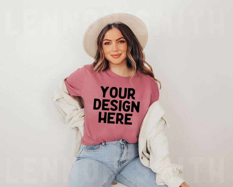 May include: A woman wearing a pink t-shirt with the text "YOUR DESIGN HERE" in black lettering. She is wearing a white hat and a white cardigan. She is sitting on a white surface.