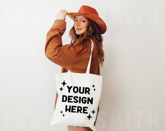 May include: A white tote bag with black text that says "YOUR DESIGN HERE" and small stars around the text. The bag is being held by a person wearing a brown jacket and a brown hat.