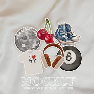 Puede incluir: Un conjunto de pegatinas que incluyen una bola de discoteca, un par de zapatillas azules, una cereza roja, unos auriculares y una bola de billar negra y blanca. Las pegatinas están sobre un fondo de tela blanca.
