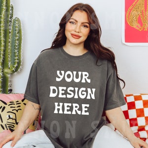 May include: A woman wearing a dark gray t-shirt with the text "YOUR DESIGN HERE" in white. She is sitting on a couch with a pink and white patterned pillow and a red and white checkered pillow behind her.