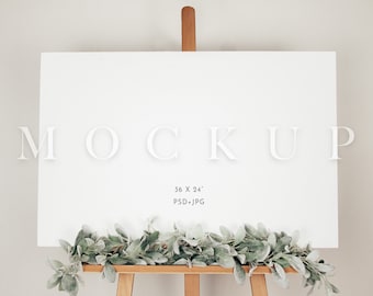 Pode incluir: Maquete de tela branca em branco em um cavalete com uma guirlanda de folhagem verde. A tela mede 91,44 x 60,96 cm e está etiquetada como "MOCKUP".
