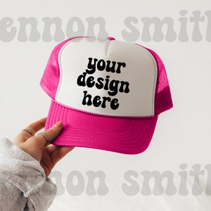 Könnte beinhalten: Eine weiße und rosafarbene Trucker-Kappe mit Netzrücken. Die Vorderseite der Kappe hat schwarzen Text mit der Aufschrift "your design here".