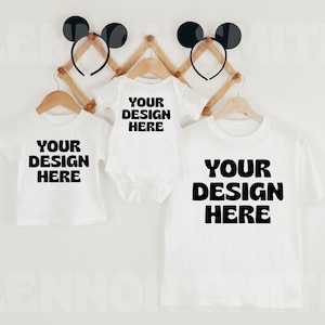 K&ouml;nnte beinhalten: Drei wei&szlig;e T-Shirts und ein Baby-Body h&auml;ngen an Holzkleiderb&uuml;geln. Jedes Kleidungsst&uuml;ck tr&auml;gt den Text "YOUR DESIGN HERE" in fetten schwarzen Buchstaben. Zwei schwarze Ohren-Stirnb&auml;nder sind &uuml;ber den Hemden angebracht.