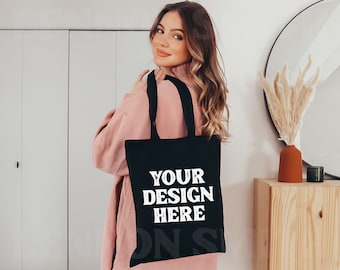 May include: A woman is holding a black tote bag with white text that says "YOUR DESIGN HERE". The bag has long black straps. The woman is wearing a pink sweater.