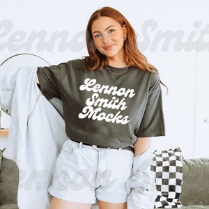 May include: A woman wearing a dark grey t-shirt with the text "Lennon Smith Mocks" in white retro font. She is wearing light blue denim shorts and a light blue denim jacket.