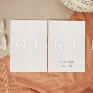 Op de afbeelding: Twee lege witte kaarten op een bruine stoffen achtergrond met een witte, pluche plant in de hoek. De kaarten zijn 5 x 7 inch en zijn gelabeld "MOCKUP 5 X 7" RATIO PSD+JPG".