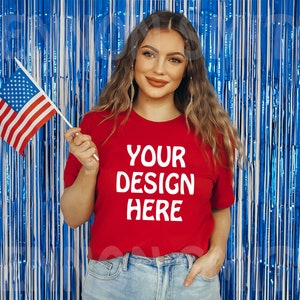 K&ouml;nnte beinhalten: Eine Frau tr&auml;gt ein rotes T-Shirt mit dem Text "YOUR DESIGN HERE" in Wei&szlig;. Sie h&auml;lt eine kleine amerikanische Flagge in ihrer rechten Hand. Der Hintergrund ist ein blauer und silberner Lametta-Vorhang.