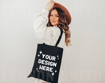 May include: A woman wearing a white sweater and a brown hat is holding a black tote bag with the text "YOUR DESIGN HERE" in white lettering and white stars.