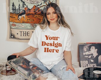 May include: A white t-shirt with the text "Your Design Here" in orange letters. The shirt is being worn by a person with long brown hair. The person is sitting on a light brown surface with a record player and a record on the surface.
