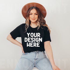 May include: A woman wearing a black t-shirt with the text "YOUR DESIGN HERE" in white, blue jeans, and a brown hat.