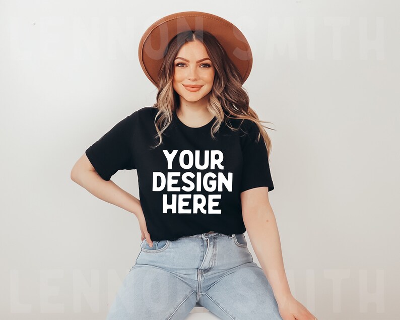 May include: A woman wearing a black t-shirt with the text "YOUR DESIGN HERE" in white, blue jeans, and a brown hat.
