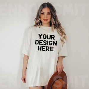Puede incluir: Una mujer lleva una camiseta blanca de gran tama&ntilde;o con el texto "YOUR DESIGN HERE" impreso en negro. Lleva un sombrero marr&oacute;n en la mano derecha.