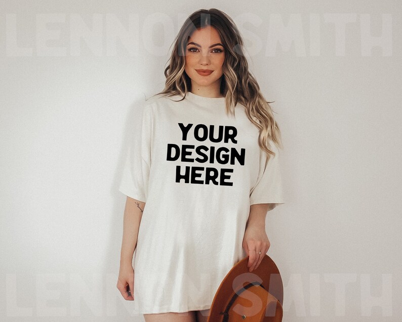 Puede incluir: Una mujer lleva una camiseta blanca de gran tama&ntilde;o con el texto "YOUR DESIGN HERE" impreso en negro. Lleva un sombrero marr&oacute;n en la mano derecha.