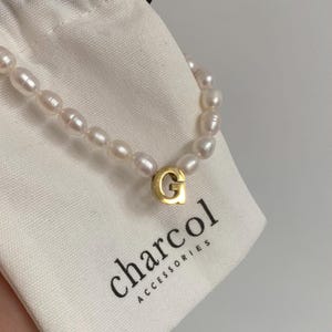 May include: A white pearl necklace with a gold letter G charm. The necklace is on a white fabric bag with the text "charcoal accessories".