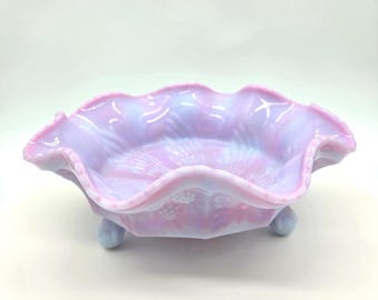 May include: A decorative, iridescent glass bowl in shades of pink and lavender. The bowl has a ruffled edge and sits on three small, rounded feet. The interior features a textured pattern, and the overall design is ornate and elegant.