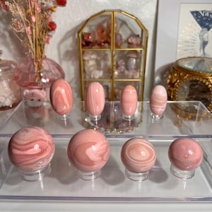 May include: A collection of pink and white gemstone spheres and oval shapes displayed on clear stands. The stones have a swirling pattern and are arranged on a clear tray. Decorative items are in the background.