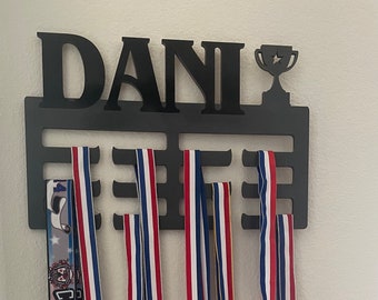 以下が含まれることがあります： 「DANI」と刻まれた黒い金属製のメダルディスプレイラック。ラックにはメダルを収納できるスロットが10個と、上部にトロフィーホルダーが付いています。ラックには赤、白、青のリボンが付いたメダルが5個掛かっています。
