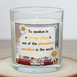 May include: A clear glass candle with a white wax interior. The candle has a label with the text "To awaken in a strange place is one of the pleasantest sensations in the world." The label also features a red and white van illustration and daisy graphics.