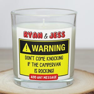 May include: A clear glass candle with a white wax interior. The candle has a label with the text "RYAN & JESS" above a yellow warning sign that reads "WARNING DON'T COME KNOCKING IF THE CAMPERVAN IS ROCKING!" and "ADD ANY MESSAGE".