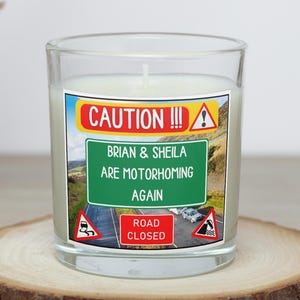 May include: A clear glass candle with a white wax interior. The candle has a label with the text "CAUTION!!!" and "BRIAN & SHEILA ARE MOTORHOMING AGAIN." The label also features road signs and a scenic background. The candle sits on a wooden slice.