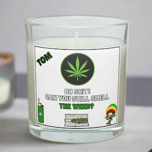 Puede incluir: Una vela de vidrio transparente con un interior de cera blanca. La vela tiene una etiqueta con el texto "OH SHIT! CAN YOU STILL SMELL THE WEED?" e ilustraciones de una hoja de marihuana, un encendedor, una bolsa de hierba y un personaje de dibujos animados.