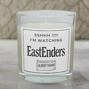 May include: A glass candle with a white label featuring the text "SSHHH !!!!! I'M WATCHING EastEnders" and a London Borough of Walford, Albert Square E20 logo. The candle is filled with white wax and sits on a light surface.