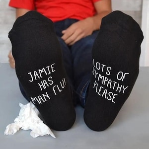 May include: Black socks with white text that reads "JAMIE HAS MAN FLU!" on one sock and "LOTS OF SYMPATHY PLEASE" on the other. A few tissues are on the floor. The socks are a humorous gift.