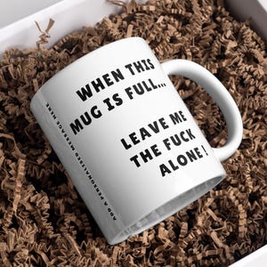 Puede incluir: Taza de cerámica blanca con texto negro: "WHEN THIS MUG IS FULL... LEAVE ME THE FUCK ALONE!". La taza está dentro de una caja blanca llena de papel arrugado marrón. La taza tiene un asa blanca.
